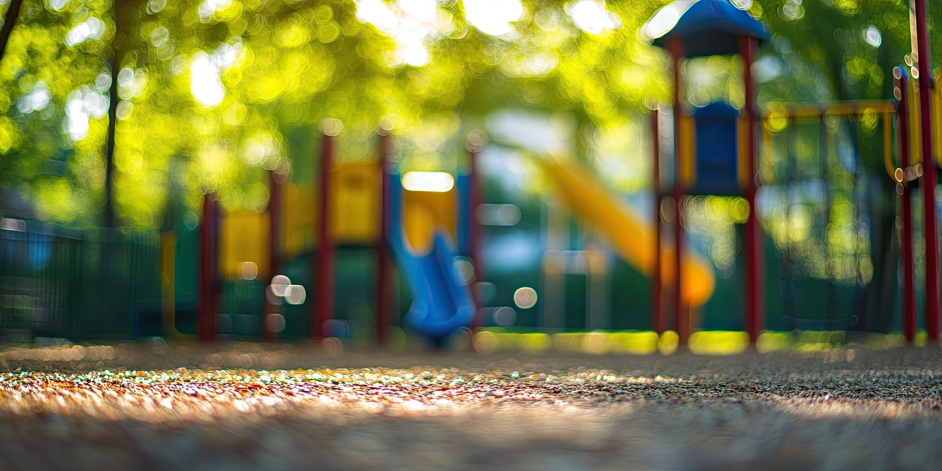Blurry playground scene in a vibrant public park with colored slides and swings softly illuminated by dappled sunlight through green foliage.