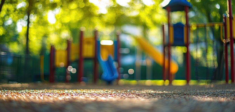 Blurry playground scene in a vibrant public park with colored slides and swings softly illuminated by dappled sunlight through green foliage.